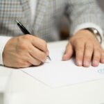 A person signing a document with a pen, with paper and a contract visible on the surface.