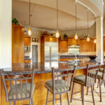 An open kitchen with a curved granite breakfast bar, metal barstools and warm wood cabinets.