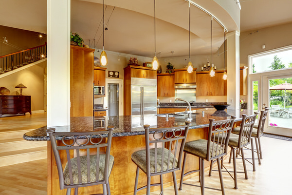 An open kitchen with a curved granite breakfast bar, metal barstools and warm wood cabinets.