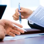 A hand holding a pen over a document on a desk beside a laptop and notebook.