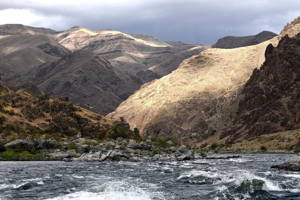 Rocky landscape along the Snake River in Hells Canyon, Idaho, with mountains, grass, trees, and river rapids.