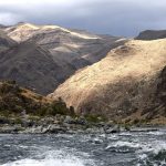 Rocky landscape along the Snake River in Hells Canyon, Idaho, with mountains, grass, trees, and river rapids.